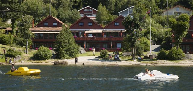 Mones Feriesenter sett fra sjøen — røde hytter, tretterrasser, sandstrand og tråbåter på Skogsfjorden.